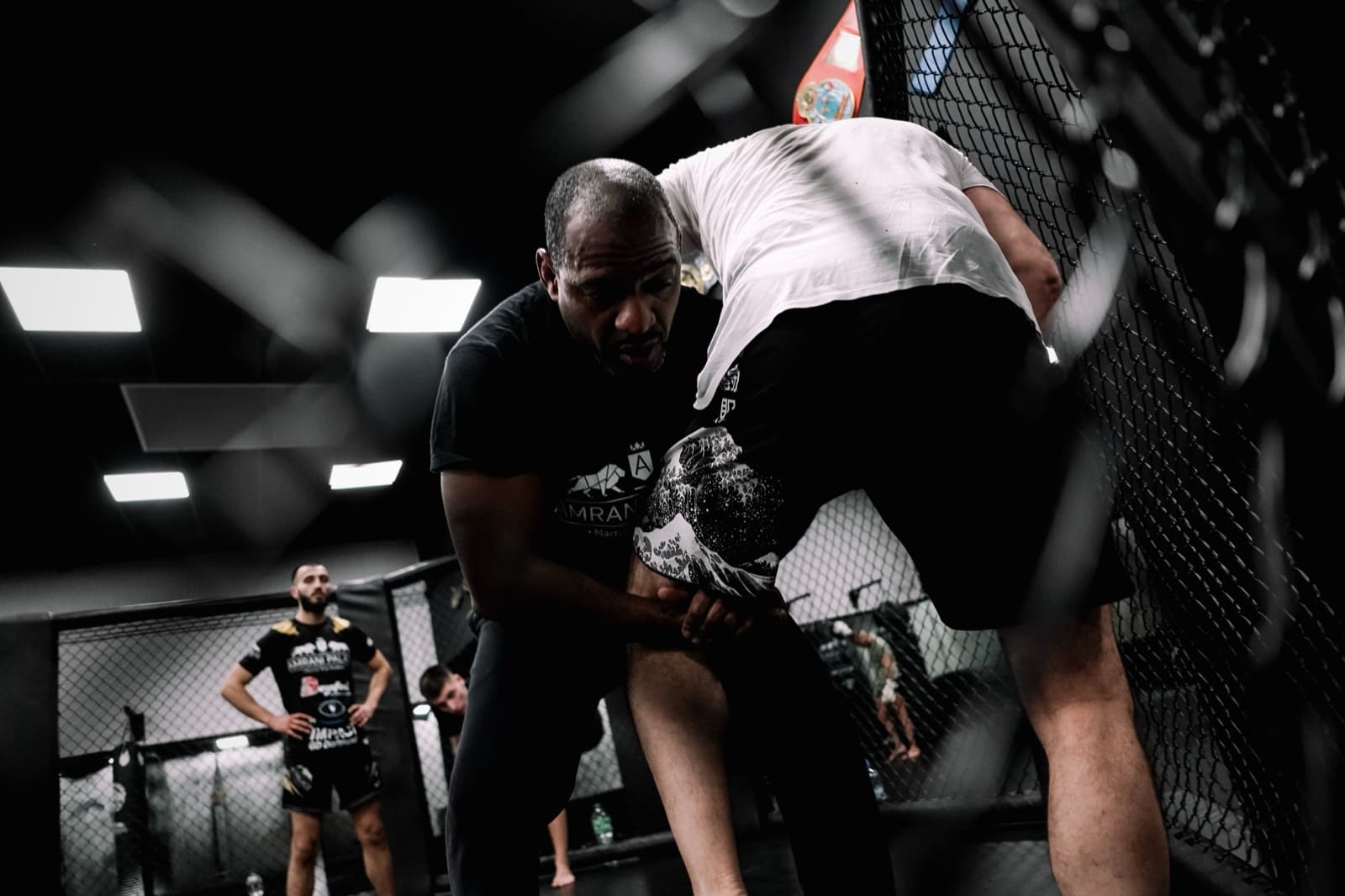 Zwei Sportler ringen im MMA-Käfig am Gitter, einer im Amrani-Palace-Shirt greift das Bein des anderen, Training im Amrani Palace Oberhausen